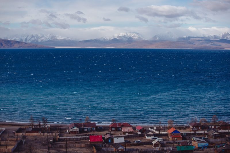 Lakeside Mongolian village with colourful rooftops and snowy mountains in the background.
