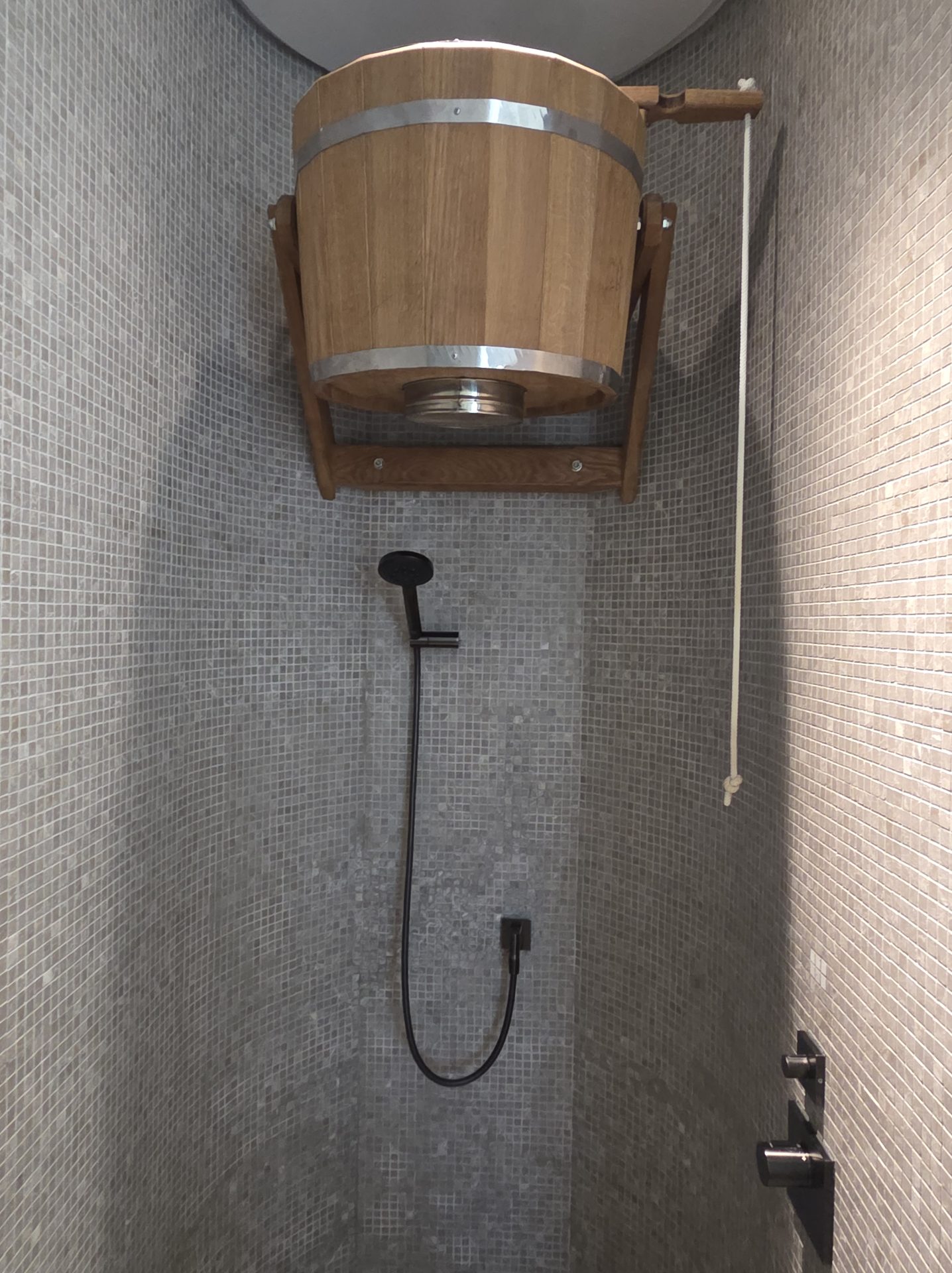Traditional Russian banya wooden dousing bucket mounted on the wall above a handheld shower in the Banya Spa House at Aman Nai Lert Bangkok, with a rope pull for releasing icy water and gray mosaic-tiled walls