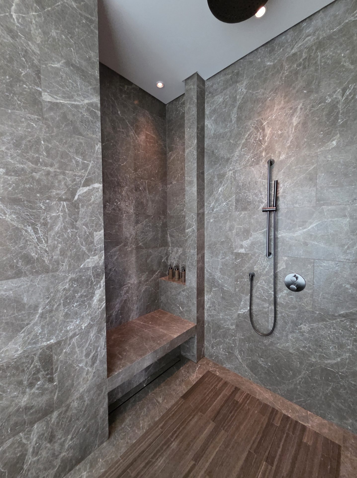 Walk-in rain shower in a Corner Suite at Aman Nai Lert Bangkok, with veined gray marble walls, dark matte black fixtures, a stone bench seat, and wide-plank oak flooring at the threshold
