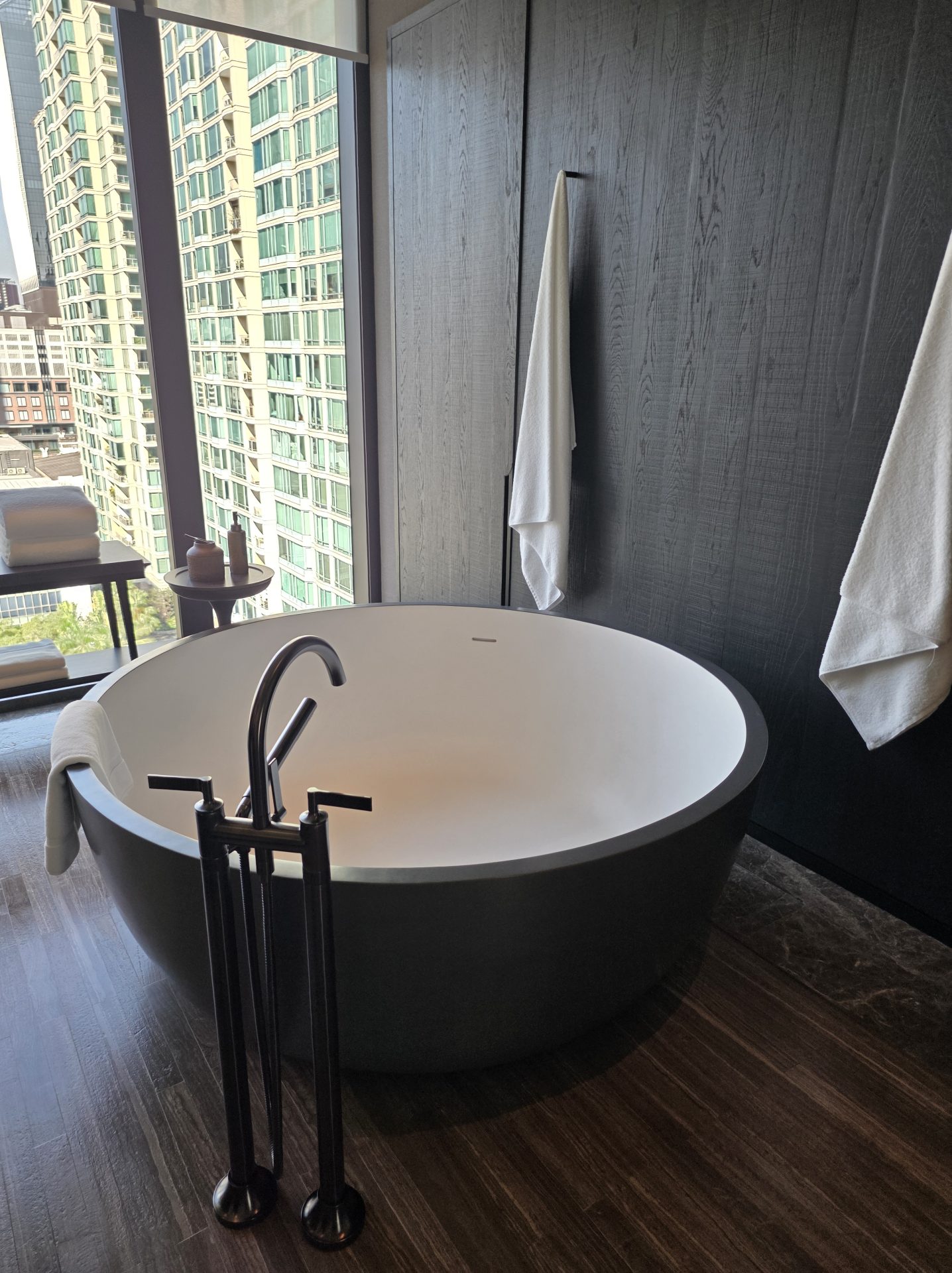 Freestanding round soaking bathtub at Aman Nai Lert Bangkok Corner Suite, white interior set into a dark stone shell with matte black floor-standing taps, positioned beside a floor-to-ceiling window with a view of a neighboring Bangkok tower