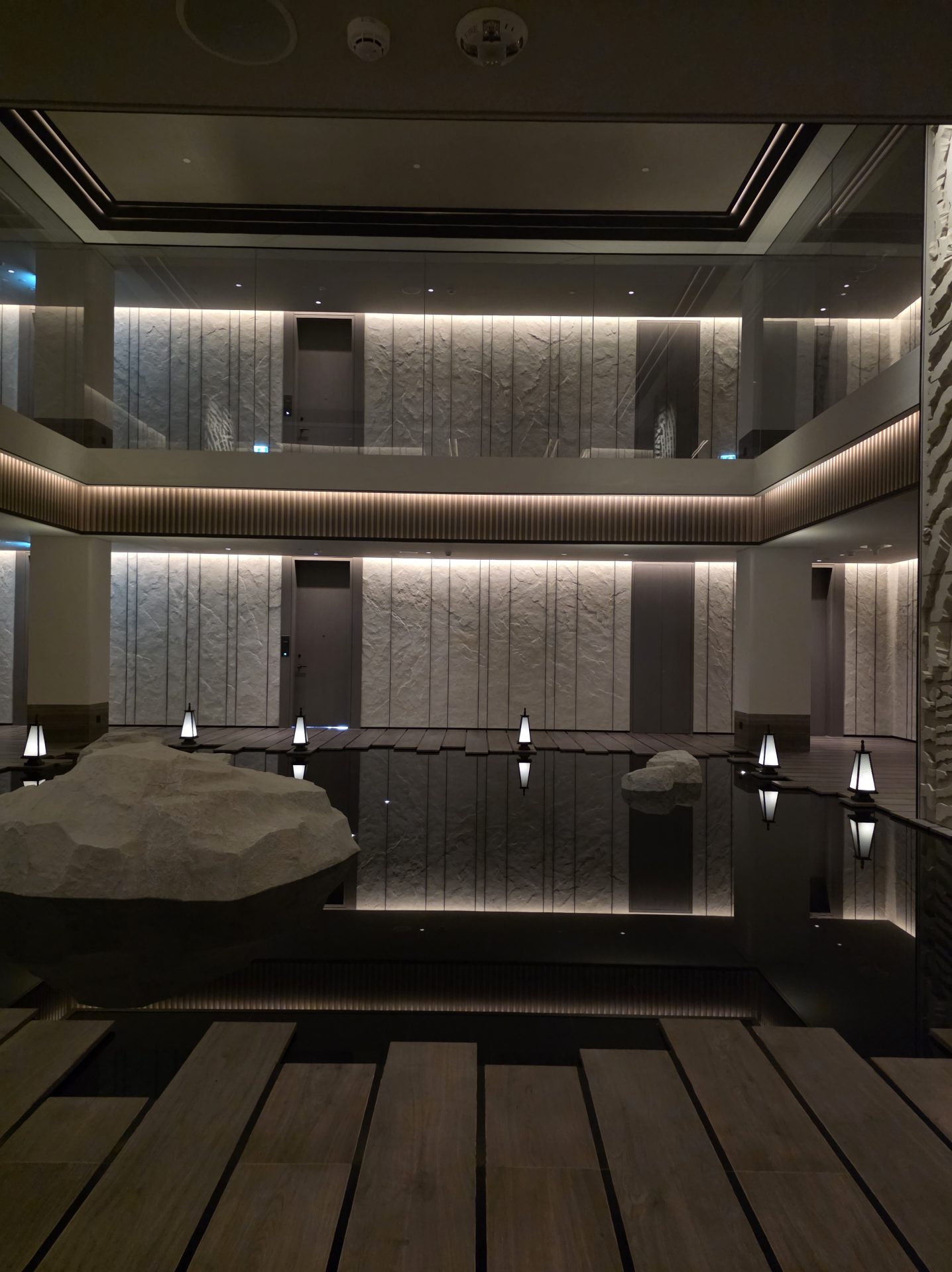 Double-height guest-floor elevator lobby at Aman Nai Lert Bangkok with a long black reflecting pool, a hand-carved white stone boulder, vertical lantern pillars, and rough-textured stone wall panels — suite doors visible at the back