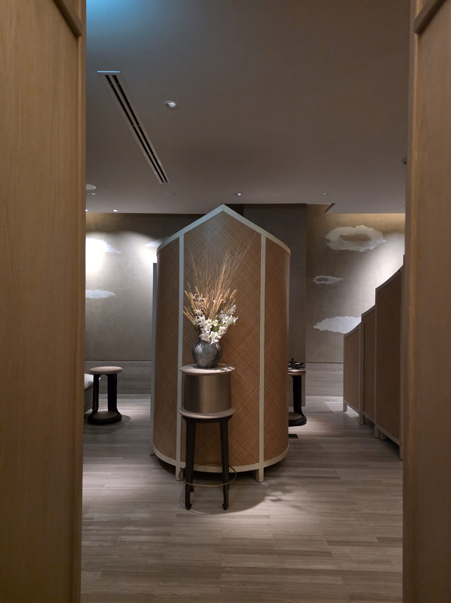 Pre-treatment relaxation and changing area at Aman Nai Lert Bangkok spa with woven cylindrical privacy screens, a tall arrangement of dried wheat and white orchids in a dark ceramic vessel, and cloud-shaped reliefs on the wall