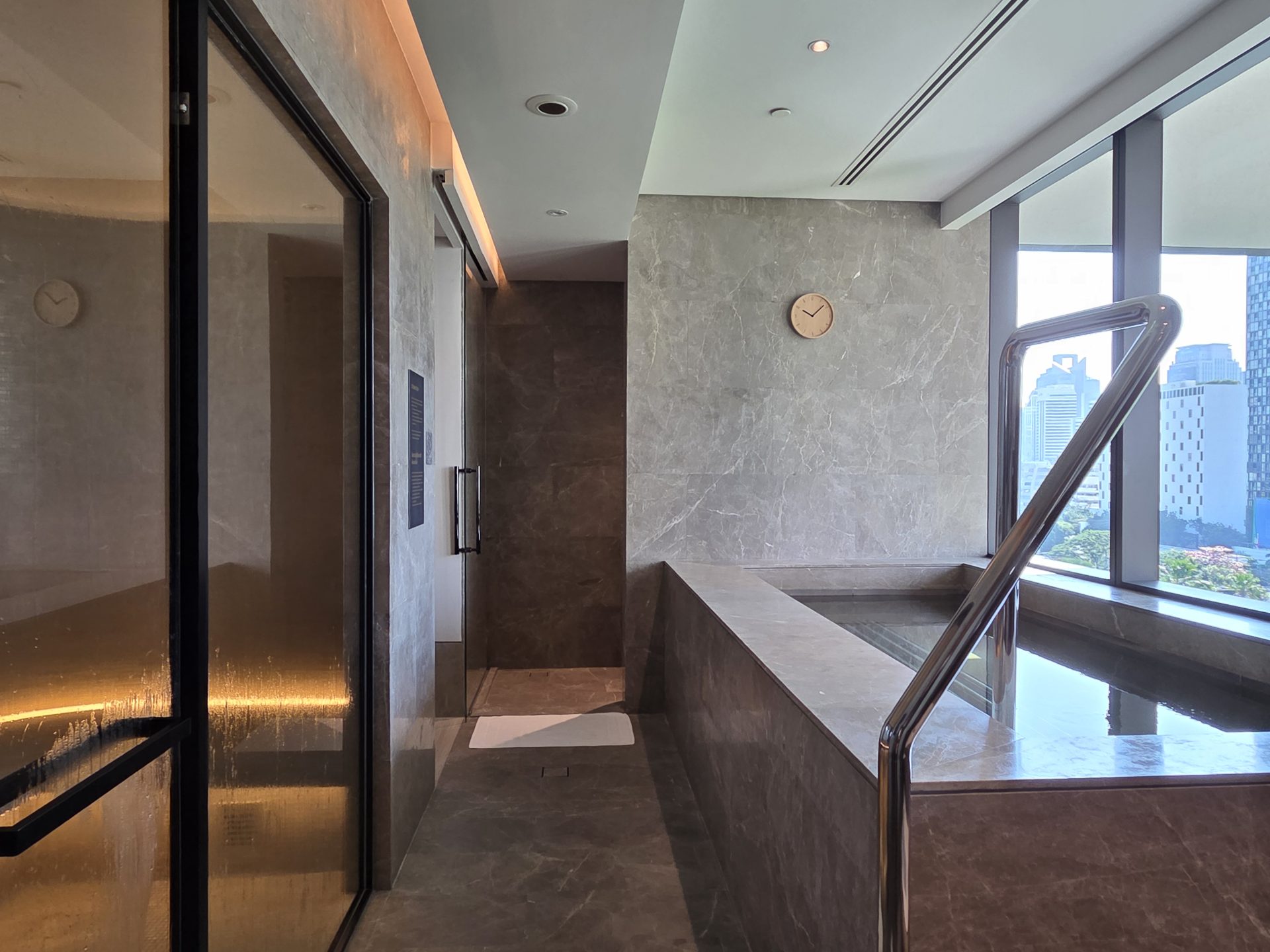 Spa wet zone at Aman Nai Lert Bangkok showing the Finnish sauna behind glass on the left and the heated marble plunge pool on the right, with a floor-to-ceiling window opening onto the Bangkok cityscape
