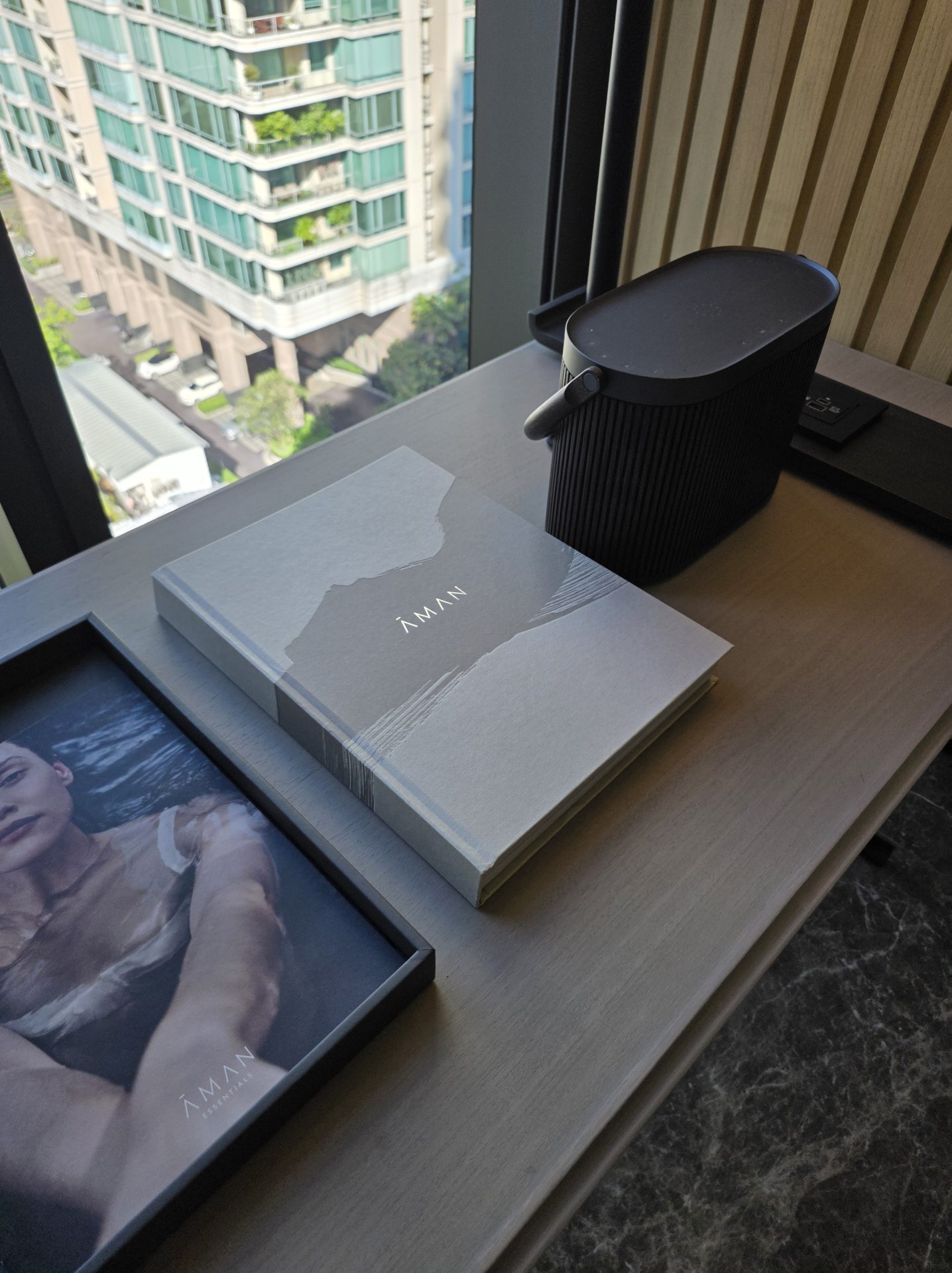 Aman welcome book and Aman Essentials grooming box on a desk at Aman Nai Lert Bangkok beside a small black ribbed speaker, with Bangkok residential buildings visible through the window