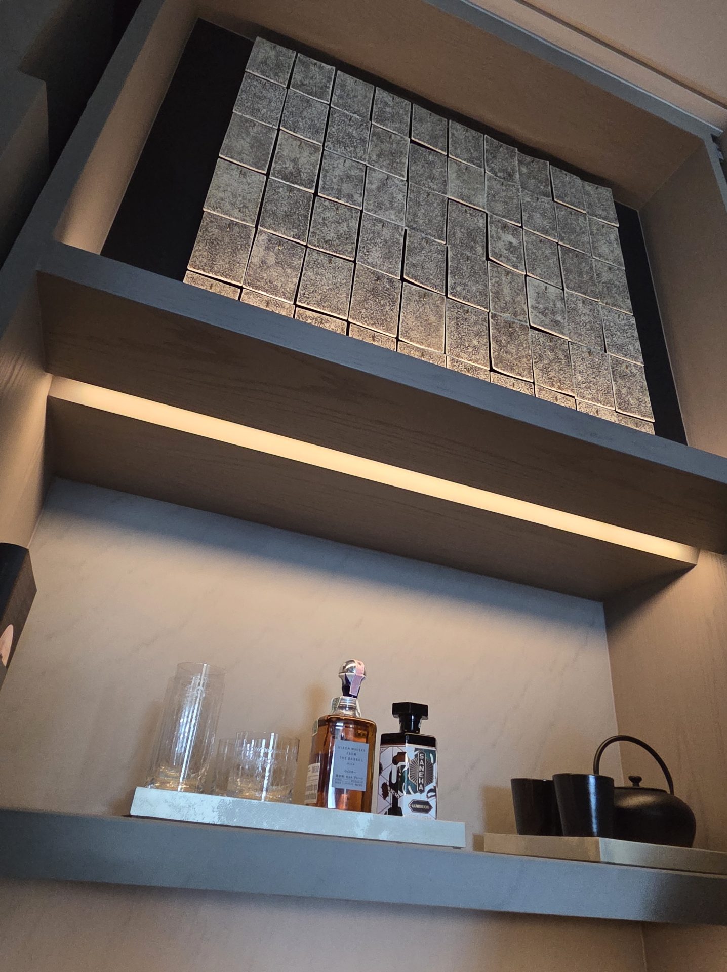 In-suite spirits display niche at Aman Nai Lert Bangkok with a backlit textured-stone wall above a shelf holding Nikka Whisky From The Barrel, a bottle of Saneha Thai liqueur, crystal glassware, and a small black ceramic teapot