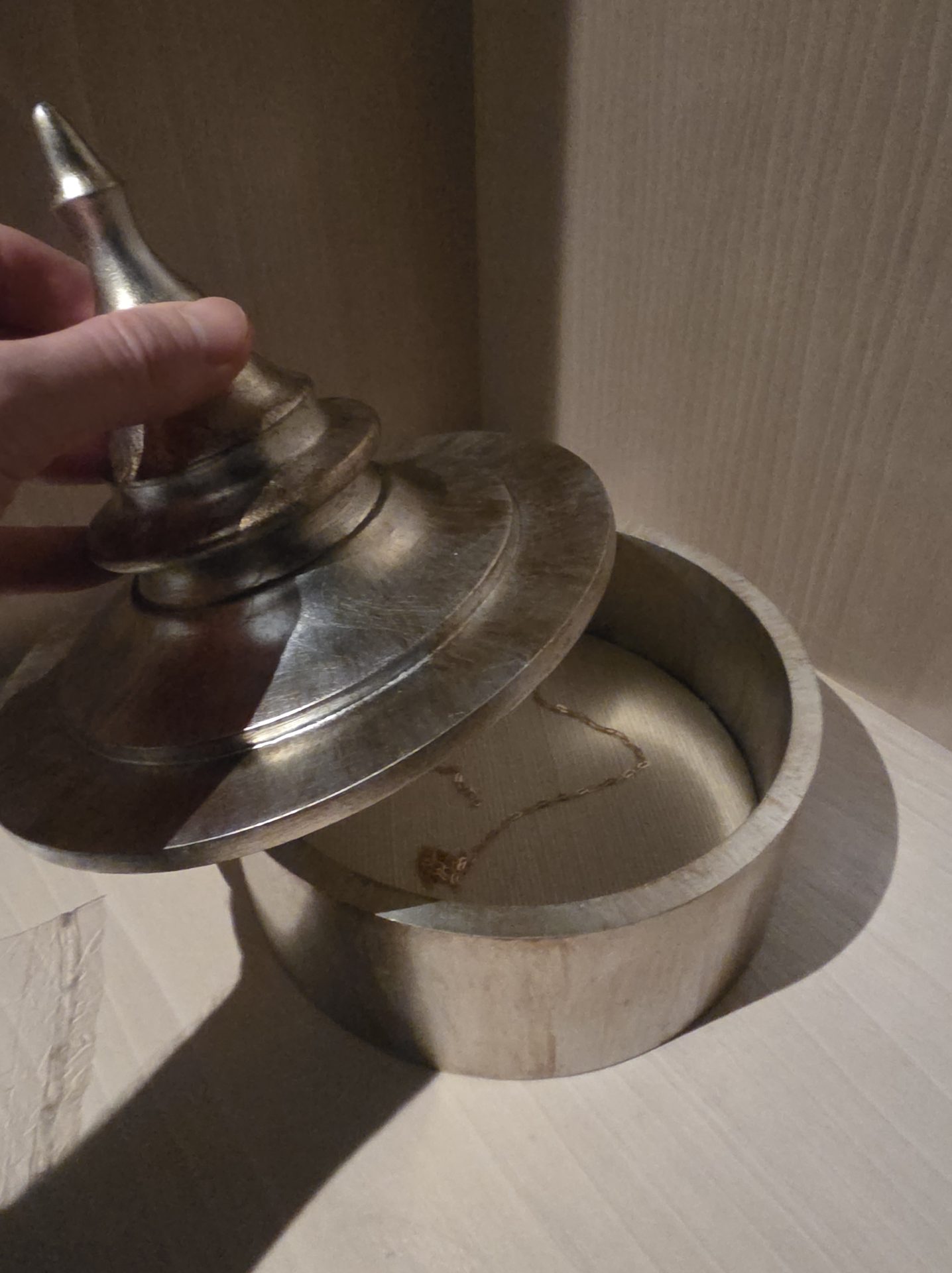 Hand opening a small carved wooden box at Aman Nai Lert Bangkok with a turned-metal lid topped by a sculptural spinning-top finial, revealing a delicate chain inside