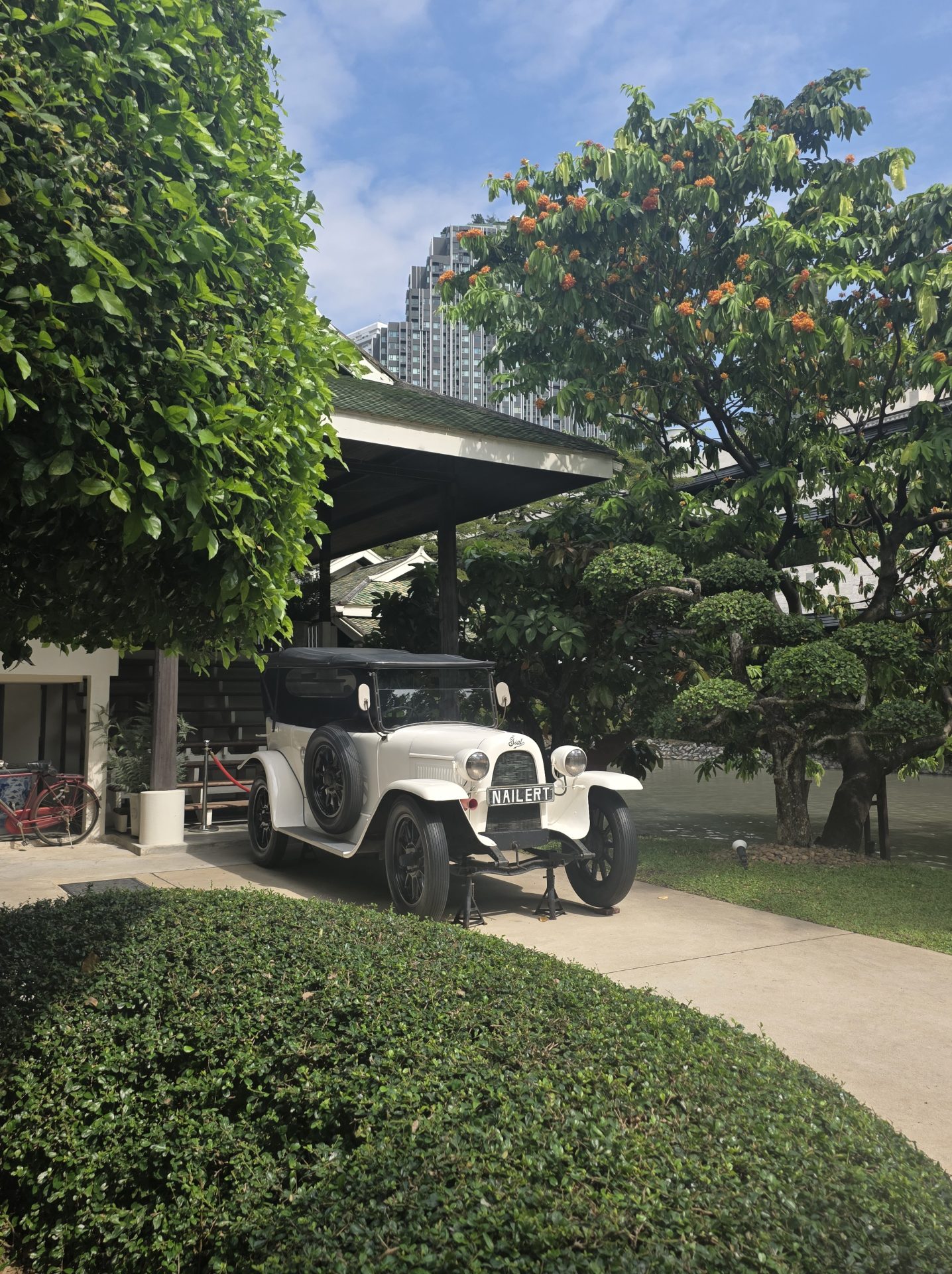 White vintage early-20th-century motor car with the "NAILERT" license plate, parked under a wooden carport at the Nai Lert Park Heritage Home in Bangkok, surrounded by tropical gardens, an orange-flowering tree, and the modern Bangkok skyline behind