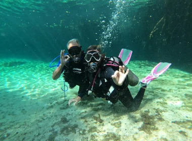 Two scuba divers posing underwater in a clear cenote near Tulum, Mexico, making the OK hand signal during a dive.