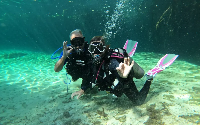 Two scuba divers posing underwater in a clear cenote near Tulum, Mexico, making the OK hand signal during a dive.
