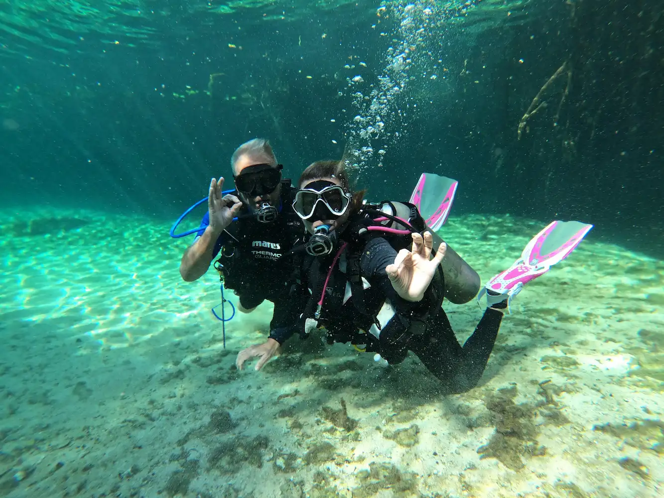 Two scuba divers posing underwater in a clear cenote near Tulum, Mexico, making the OK hand signal during a dive.
