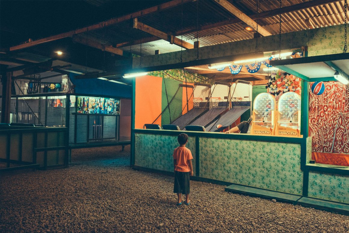 A young boy stands in front of an arcade game booth at night in French Polynesia. Photo by Hélène Havard.