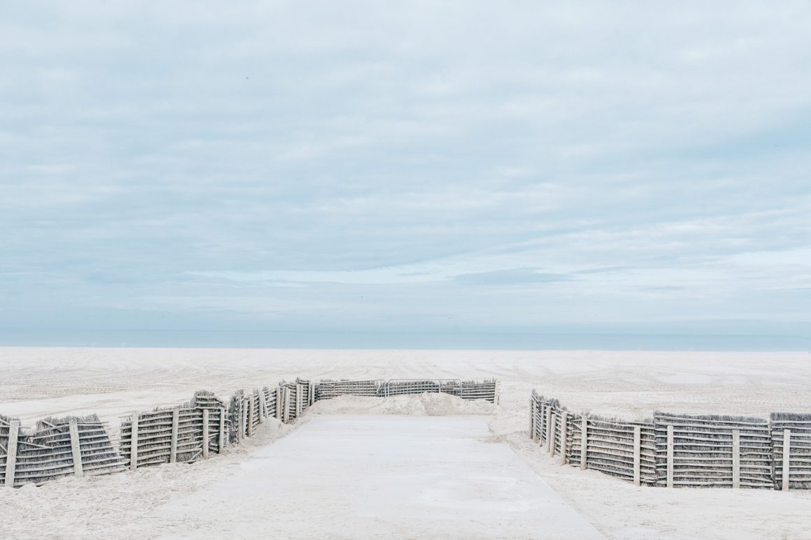 A quiet sandy path leads through beach fencing toward the sea in Normandy, France. Photo by Hélène Havard.