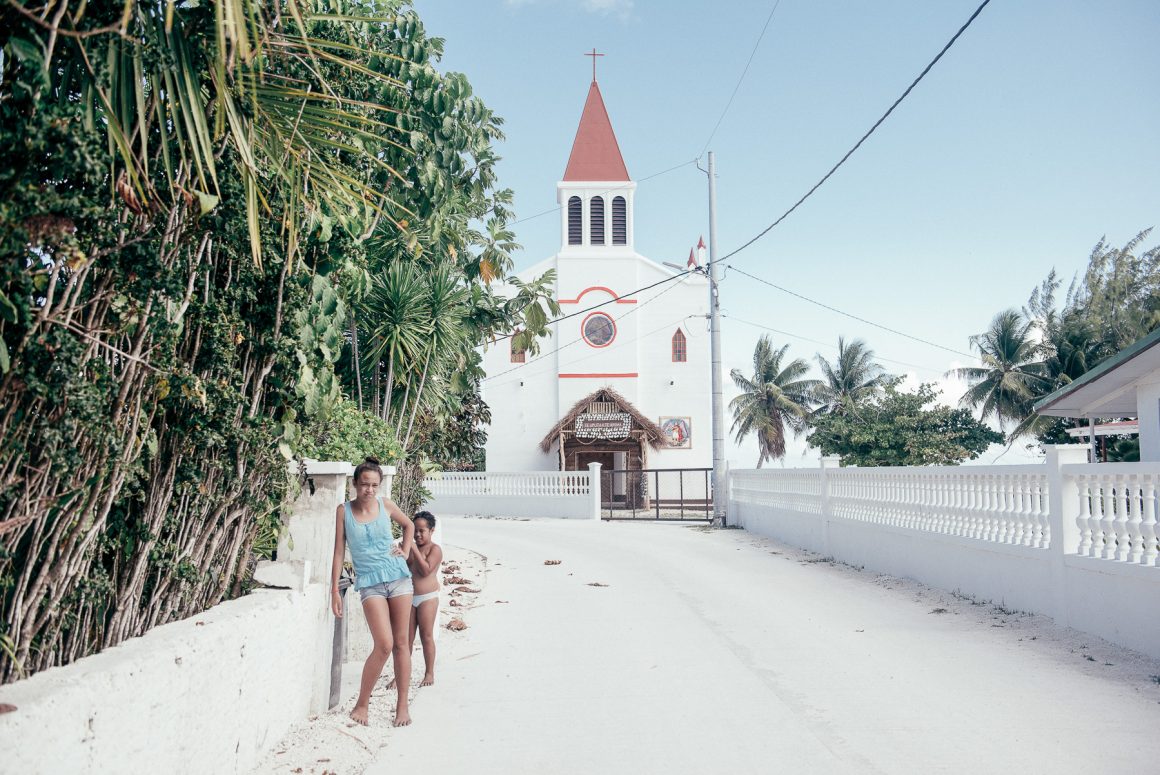 A woman and child walk past a white church on a tropical island in French Polynesia. Photo by Hélène Havard.
