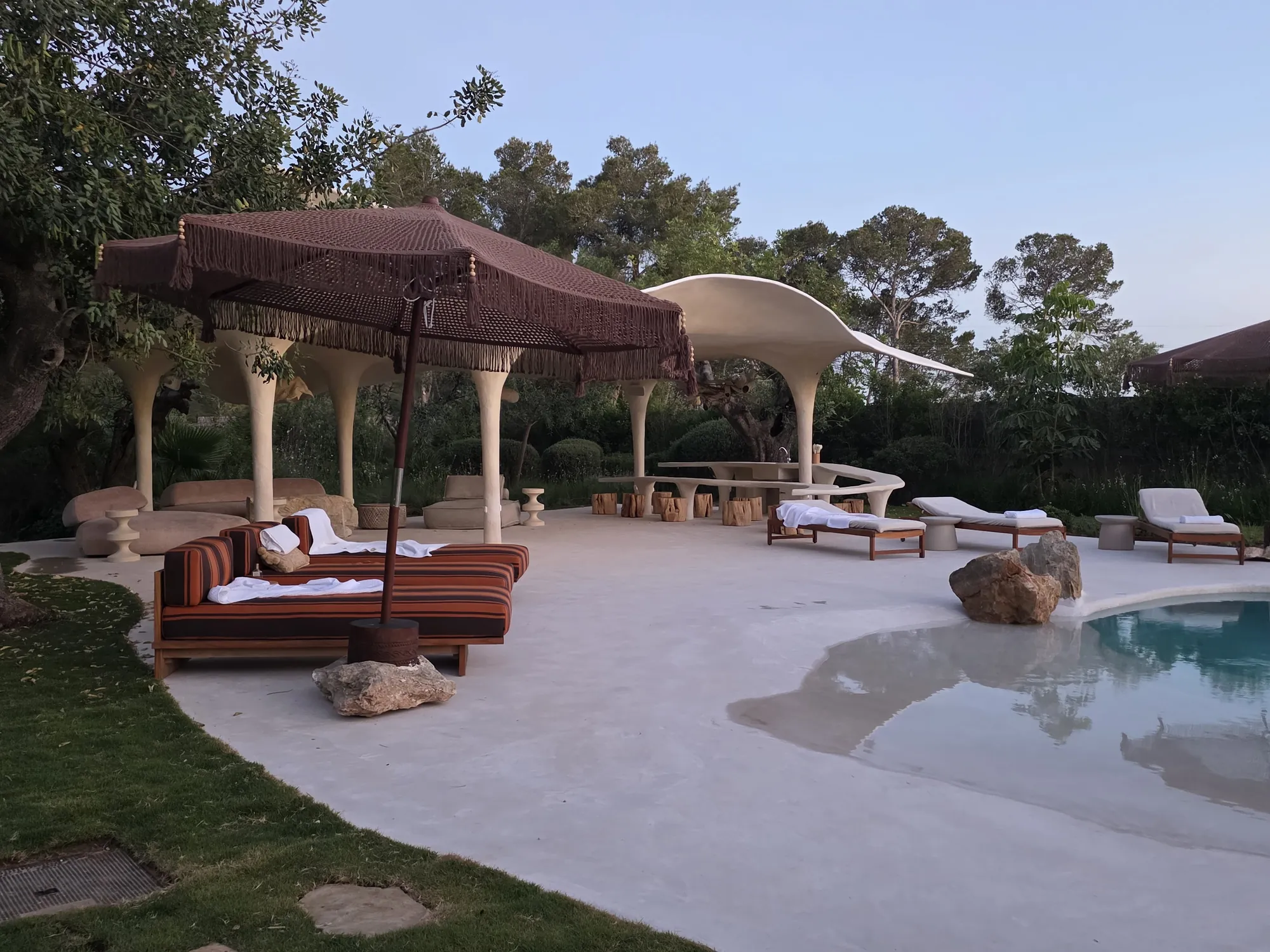 Boutique hotel pool at dusk with striped daybeds under a fringed parasol and a sculptural white pavilion