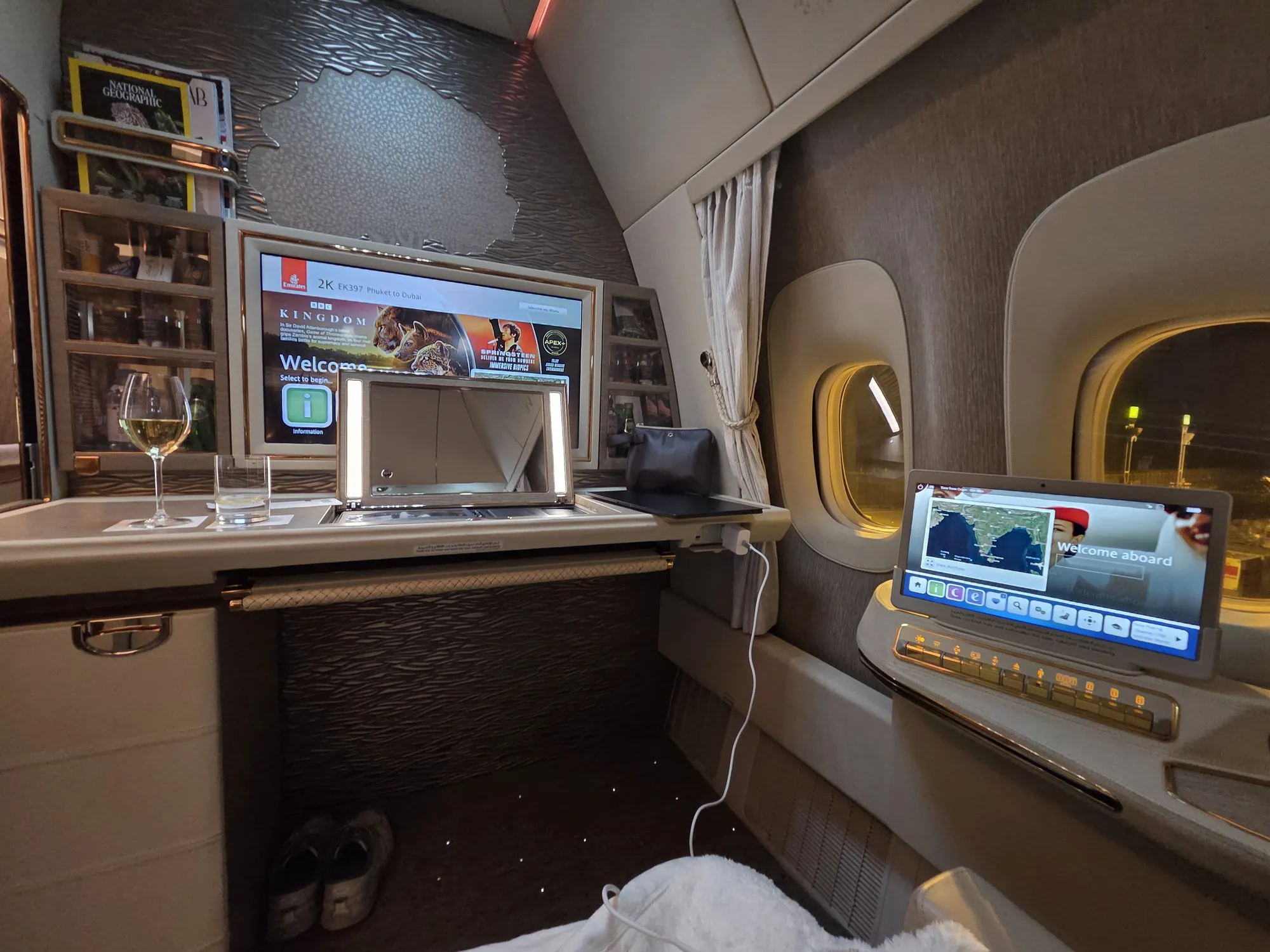 Emirates first class suite with a glass of white wine, large screen and lit window before departure