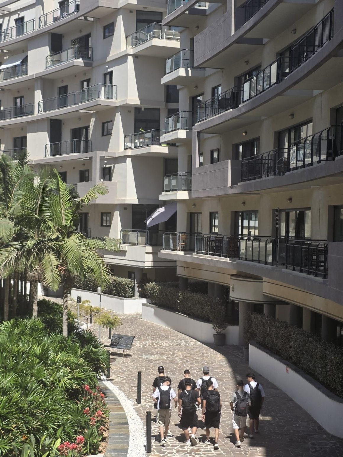 A group of young travelers with backpacks walking through the landscaped courtyard of a gated apartment complex in Malta