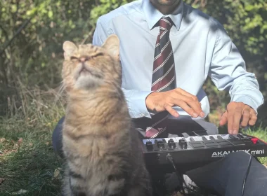 Fery Lionfarsi plays a MIDI keyboard in a blue shirt and striped tie as his tabby cat sits beside him with its eyes closed