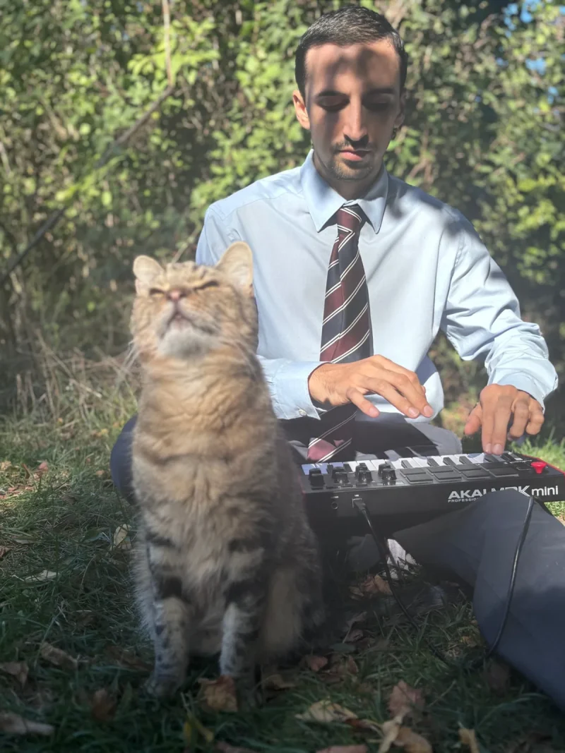 Fery Lionfarsi plays a MIDI keyboard in a blue shirt and striped tie as his tabby cat sits beside him with its eyes closed